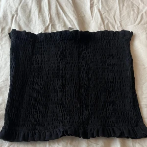 Svart bandeau tubtopp från H&M - Supersöt svart bandeau tubtopp från H&M med smockad struktur och volangkant upptill och nedtill. Perfekt att styla med höga jeans eller kjol för en trendig look. Toppens material är mjukt och stretchigt för en bekväm passform.
