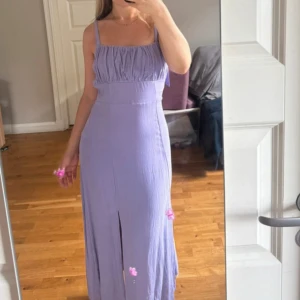 Lila långklänning med knytband NA-KD - Supersöt lila långklänning från NA-KD x Anna Briand med smockad överdel, knytband på axlarna och slits framtill. Klänningen har en flowy passform och raka axelband, perfekt för sommarens alla tillfällen. Diskret dragkedja vid sidan som gör det enkelt att få på och av sig klänningen. Aldrig använd.