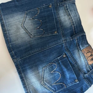 Blå jeanskjol från Monday Denim - Snygg blå jeanskjol från Monday Denim med slitningar och coola detaljer på bakfickorna, många av kristallerna har dock ramlat av. Dubbla knappar i midjan och markerade sömmar. Perfekt för dig som gillar en avslappnad och trendig stil.