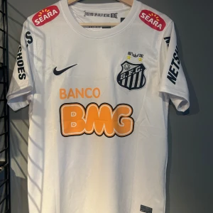 Vit Santos Neymar Jr fotbollströja Nike - Santos FC matchtröja i vitt med Neymar Jr och nummer 11 på ryggen. Tröjan har korta ärmar, flera sponsorloggor och klubbmärke på bröstet. Tillverkad i lätt och ventilerande material, perfekt för fotbollsälskare som vill sticka ut på planen. Helt ny med tagg en replika 