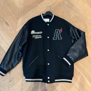 Svart bomberjacka från Zara med brodyr - Svart bomberjacka från Zara med coola broderade detaljer som 'Dream now', 'Arizona Roadtrip' och ett stort A med vinge. Passar XS. Jackan har svarta ärmar i skinnimitation, ribbade muddar i svart och beige samt knappar framtill. Perfekt för dig som vill sticka ut.