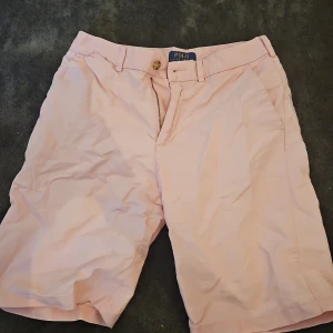 Ljusrosa shorts från Polo Ralph Lauren - Snygga ljusrosa shorts från Polo Ralph Lauren med klassisk passform. De har dragkedja, knappstängning och bälteshällor. Bakfickor med knappar och broderad grön logga på ena fickan. Perfekta för sommardagar och chill häng.