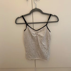 Randig linnetopp med spetsdetalj - Säljer en vit och svart randig linnetopp med tunna axelband och söt spetskant vid ringningen. Toppen har en liten rynkdetalj framtill som ger extra stilpoäng. Perfekt för varma dagar eller som layering-piece.