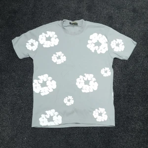 Grå blommig t-shirt från Denim Tears - Snygg grå t-shirt från Denim Tears med vita blommiga tryck över hela plagget. Klassisk passform med rund hals och korta ärmar. Perfekt för dig som vill sticka ut med en unik och trendig stil.
