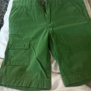 Gröna Sail Racing shorts med fickor - Snygga gröna shorts från Sail Racing med flera praktiska fickor, bland annat en stor benficka med lock. Klassisk passform och bälteshällor. Perfekta för sommardagar och äventyr. Tydlig logga baktill och detaljer i mörkblått på insidan av linningen.