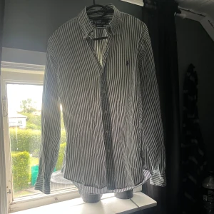 Randig skjorta från Polo Ralph Lauren - Snygg skjorta från Polo Ralph Lauren i vitt och mörkgrönt med smala vertikala ränder. Custom fit-modell med klassisk krage, långa ärmar och knäppning framtill. Broderad logga på bröstet. Perfekt för dig som gillar stilrena och tidlösa plagg.