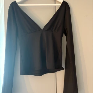 Svart långärmad topp med knytning i bak från Nelly - Snygg svart långärmad topp med djup v-ringning och stor rosett i ryggen. Toppens material känns mjukt och stretchigt, perfekt för att skapa en cool och stilren look. Passformen är normal och ärmarna är långa för en elegant vibe. Använd en gång