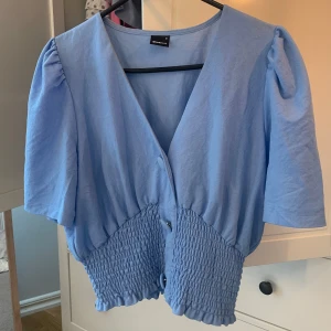 Ljusblå blus från Gina Tricot - Supersöt ljusblå blus från Gina Tricot med v-ringning, puffiga korta ärmar och smockad midja. Blusen har knappar framtill och en luftig, lite loose passform. Perfekt för dig som gillar en feminin och trendig look.