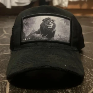 Svart keps från LAVY med lejonmotiv - Svart keps från LAVY med mesh på sidorna och baksidan för extra ventilation. Framtill finns en stor patch med ett detaljerat svartvitt lejonmotiv. Kepsen har böjd skärm och justerbar passform. Perfekt för dig som vill sticka ut med ett djurmotiv.