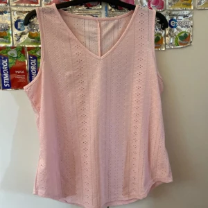 Ljusrosa linne  - Söt ljusrosa ärmlös linnetopp med v-ringning och broderat hålmönster. Toppen är tillverkad i mjuk polyesterblandning med lite stretch för extra komfort. Perfekt för varma dagar när du vill ha en luftig och stilren look.