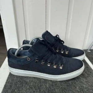 Mörkblå sneakers med mockadetaljer - Snygga mörkblå sneakers med ovandel i textil och mockadetaljer. Skorna har vita platta sulor och coola metallöglor för snörningen som ger en sportig känsla. Perfekta för dig som gillar stilrena och moderna skor med en twist.