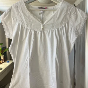 Vit blus med spets från Hillevi - Supersöt vit blus från Hillevi med korta ärmar, spetsdetaljer längs halsringningen och ärmsluten samt små knappar framtill.💕