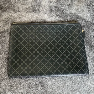 Svart datorfodral från By Malene Birger - Stilren svart datorfodral från By Malene Birger med klassiskt rutigt mönster och guldfärgad dragkedja. Datorfodralet har en svart/grå utsidaoch en ljusrosa insida med innerficka. Perfekt för laptop eller dokument. Mått: H 26,5 x B 36,5