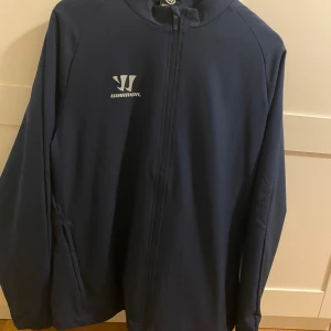 Mörkblå vindjacka från Warrior - Snygg mörkblå vindjacka från Warrior med dragkedja framtill och diskret logga på bröstet. Jackan har lång ärm och är tillverkad i ett lätt och smidigt material, perfekt för dig som gillar sportig stil.