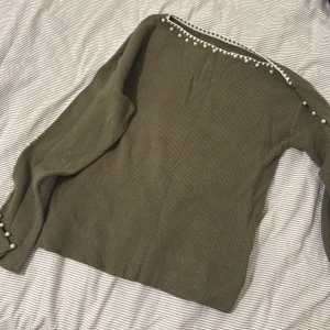 Olivgrön stickad tröja med pärlor - Olivgrön stickad tröja med långa ärmar och snygga pärldetaljer längs axlar och ärmar. Tröjan har en rak passform och ribbad struktur som ger en stilren look. Perfekt för dig som gillar unika detaljer och vill sticka ut lite extra.