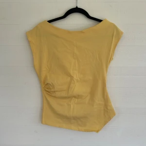 Gul ärmlös off-sholder topp från Zara - Snygg gul topp från Zara i ärmlös modell med bred halsringning och asymmetrisk nederkant. Tillverkad i mjuk bomull som känns skön mot huden. Perfekt för dig som gillar stilrena och enkla plagg med en twist. Tyvärr en liten fläck som inte går bort på framsidan av tröjan men ingenting man ser när man har den på sig då tröjan veckar så där☺️ kan skicka fler bilder vid önskemål. 