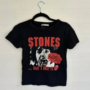Rolling Stones svart t-shirt - Svart t-shirt med Rolling Stones-tryck i rött och vitt framtill. Klassisk rund hals och korta ärmar. Materialet är mjuk bomull och passformen är normal. Perfekt för dig som gillar bandmerch och vill ha en cool vibe.