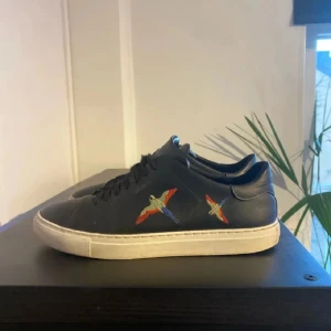Svarta Axel Arigato sneakers med broderad fågel - Svarta sneakers från Axel Arigato i skinn med broderade färgglada fåglar på sidan. Klassisk vit sula och svarta snören. Snygg och stilren modell som passar till det mesta. Broderidetaljerna ger skorna en unik touch.Litte smutsiga men kan göra rent dom inan ja fraktar.