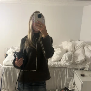 Mörkbrun stickad tröja med vit kant - Säljer en mörkbrun stickad tröja med raka, långa ärmar och en unik vit kant som går längs framsidan och ärmslut. Tröjan har en något boxig passform och hög hals, perfekt för dig som gillar stilrena detaljer och vill ha något som sticker ut.