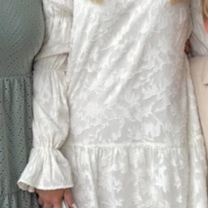 Vit blommig kortklänning med volangärm - Superfin vit kortklänning med diskret blommigt mönster och långa volangärmar. Klänningen har rund halsringning och en lätt utsvängd kjol. Perfekt för sommarens alla tillfällen och ger en romantisk vibe.