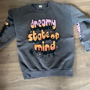 Grå med tryck - Grå sweatshirt från Lindex med färgglatt tryck 'dreamy state of mind' i lila och orange på bröstet. Blommor och retrovibbar på både framsidan och ärmarna. Mjuk insida och ribbade muddar. Perfekt för dig som gillar unika prints och comfy stil. Är lite sliten i ena ärmen men i annat bra skick 🌸