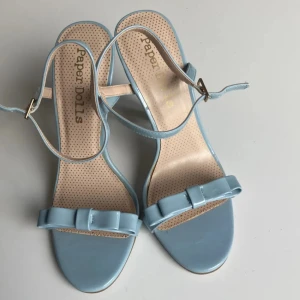 Ljusblå sandaler med rosett från Paper Dolls - Snygga ljusblå sandaler från Paper Dolls med lackad finish, smal rem runt vristen och söt rosettdetalj framtill. Skorna har öppen tå, hög klack och rundad sula. Perfekta för dig som vill sticka ut med en feminin och trendig look.