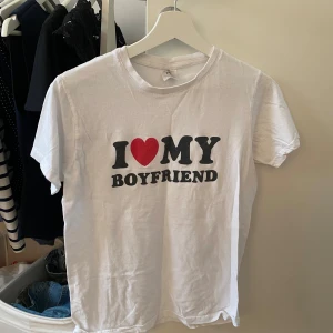 Vit t-shirt med I ❤️ MY BOYFRIEND-tryck - Vit t-shirt med klassisk passform och rund hals. Framsidan har ett stort tryck med texten 'I ❤️ MY BOYFRIEND' i svart och rött. Perfekt statement-plagg för dig som vill visa lite extra kärlek. Skön och enkel stil som funkar till jeans eller shorts.