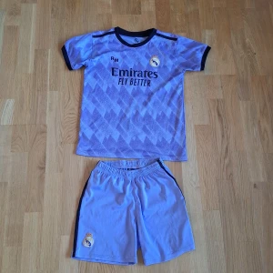 Real Madrid Benzema matchställ strl 12 "145-160" - Säljer ett Real Madrid matchställ med Benzema 9 på ryggen. Tröjan är ljuslila med mörkblå detaljer, korta ärmar och geometriskt mönster. Byxorna är ljuslila med klubbmärke. Materialet är lätt och andas, perfekt för fotbollsträning eller match.