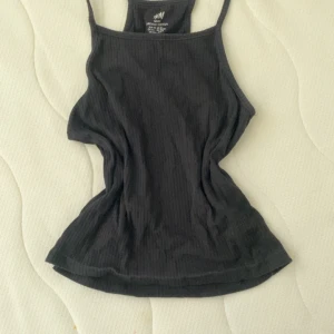 Svart ribbad linnetopp från H&M - Svart ribbad linnetopp från H&M i ekologisk bomull. Toppen har smala axelband och en rak halsringning, perfekt för en enkel och clean look. Materialet är mjukt och stretchigt för en bekväm passform.