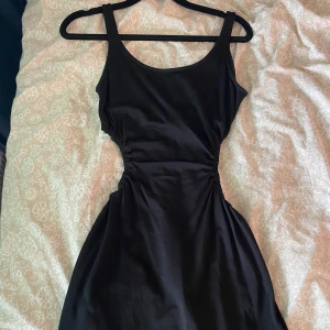 Svart tight kortklänning med cut-out detaljer - Säljer en svart tight kortklänning med rund halsringning och snygga cut-out detaljer i midjan. Klänningen är ärmlös och har en enkel, stilren design som passar perfekt till sommarens alla tillfällen. Nypris 690 kr.