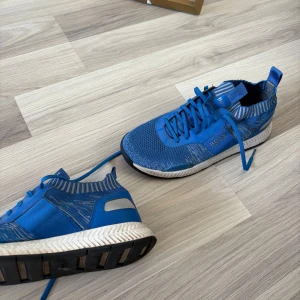 Blå sneakers från BOSS med stickad ovandel - Snygga blå sneakers från BOSS med stickad ovandel och coola mönsterdetaljer i grått och vitt. Skorna har snörning, vit sula med svarta detaljer och en sportig vibe. Perfekta för dig som gillar stilrena men ändå unika sneakers.