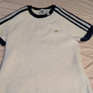 Vit teddy t-shirt från Adidas - Vit teddy t-shirt från Adidas med marinblå detaljer och klassiska tre ränder på ärmarna. Guldig Adidas-logga på bröstet. Mjuk och fluffig känsla, perfekt för dig som gillar sportig och unik stil.