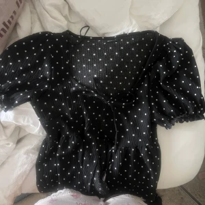 Svart prickig blus från Gina Tricot - Supersöt svart blus med vita prickar från Gina Tricot. Modellen har kort puffärm, v-ringning och knyt framtill. Tunt, lätt material med diskret ribbad struktur och volangkant nedtill. Perfekt för dig som gillar en lekfull och trendig look.