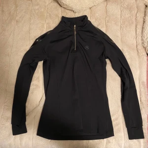 Svart långärmad tröja Alequi half zip - Svart långärmad tröja från Alequi med half zip och hög krage. Slimmad passform och diskreta loggor på bröstet, ryggen och ena ärmen. Perfekt för ridning eller träning, tillverkad i stretchigt syntetmaterial som andas.