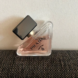 Prada Paradoxe Eau de Parfum - Lyxig parfym från Prada med en unik trekantig glasflaska och ljusrosa doftvätska. Flaskan har en stor svart rektangulär kork och eleganta detaljer med Pradas logga. Perfekt för dig som gillar exklusiva och trendiga dofter.