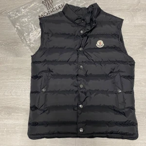 Moncler väst  - Svart dunväst med klassisk ståkrage och horisontella quiltade sömmar. Västen har tryckknappar framtill, två fickor med knappstängning och Monclers ikoniska logga på bröstet. Perfekt för lager-på-lager och snygg till både hoodie och t-shirt.