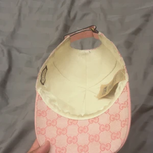 Rosa Gucci keps med GG-mönster - Snygg rosa keps från Gucci med klassiskt GG-mönster i ljusrosa och rosa toner. Justerbart band bak i rosa läder med guldfärgad spänne. Kepsen är tillverkad i bomull och har en liten guldfärgad GG-detalj på sidan. Perfekt för dig som gillar exklusiva accessoarer.