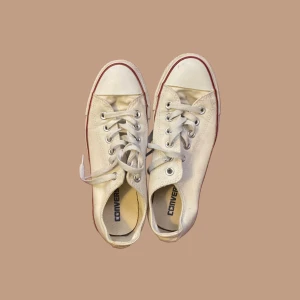 Vita Converse All Star sneakers - Klassiska vita Converse All Star sneakers med lågt skaft, röd och blå rand runt sulan och snörning framtill. Skorna har rund tå och är tillverkade i canvas med gummisula. Perfekta för en avslappnad och cool stil.