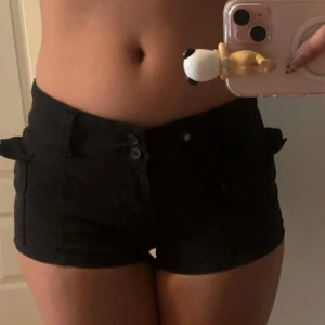 Svarta lowwaist shorts med fickor - Snygga svarta shorts me low midja och kort passform. De har praktiska fickor på sidorna och bälteshällor. Stängs med dragkedja och dubbla knappar framtill. Perfekta för varma dagar och en chill look.