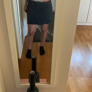Svart volang jeanskjol  - Säljer en svart kort jeanskjol med volangdetalj längst ner. Kjolen har klassisk femficksdesign, dragkedja och knapp framtill samt coola sömdetaljer framtill. Perfekt för dig som gillar en edgy och trendig look.