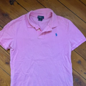 Rosa pikétröja från Ralph Lauren - Säljer en superfin rosa pikétröja från Polo Ralph Lauren med blå broderad logga på bröstet. Tröjan har korta ärmar, krage och knäppning med två knappar. Tillverkad i mjuk bomull som är skön mot huden. Storlek L men är i barnstorlek. Aldrig använd💕
