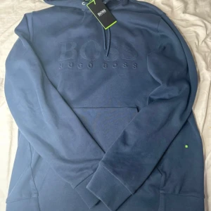 Blå hoodie från Hugo Boss med logga - Snygg marinblå hoodie från Hugo Boss med broderad logga på bröstet. Tröjan har huva med dragsko, magficka och ribbade muddar. Tillverkad i mjuk bomullsmix som känns riktigt skön mot huden. Perfekt för dig som gillar stilrena och klassiska plagg.