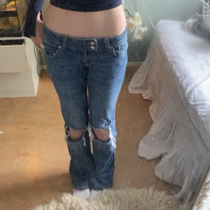 Blå bootcut jeans från 157, S - Säljer ett par blå bootcut jeans från 157 i storlek S. Jeansen har låg midja, slitna hål över knäna och klassisk femficksdesign. De är tillverkade i jeansmaterial och har dubbla knappar i midjan för extra detalj.