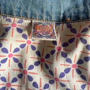 Jeansjacka från Oilily med pärlbroderi - Snygg ljusblå jeansjacka från Oilily med unika pärlbroderade blommor framtill och på ena sidan. Fodret har ett färgglatt retroinspirerat mönster i lila och rosa. Klassisk passform med knappar och bröstficka. Perfekt för dig som vill sticka ut med en kreativ vibe.