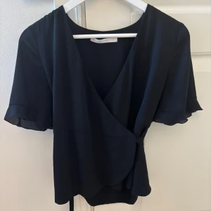 Svart omlottblus - Svart omlottblus med korta volangärmar och knyt i sidan. Blusen har en djup v-ringning och ett lätt, luftigt tyg. Storlek M men passar som S också.