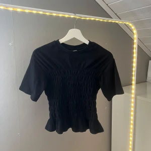 Svart smockad topp Gina Tricot - Supersnygg svart topp från Gina Tricot med smockad midja och volangkant nertill. T-shirtrund hals, korta ärmar och croppad passform. 