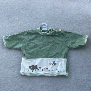 Grön kortärmad bomullströja med brodyr - Söt grön kortärmad tröja i 100% bomull med randig krage och knappar på axeln. Nedtill finns en vit panel med broderade svampar, löv, björn och fåglar. Perfekt för dig som gillar gulliga detaljer och naturinspirerat motiv.