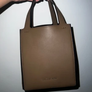 Brun handväska från Michael Kors - Jordi Small Leather Tote Bag. Snygg och stilren brun handväska från Michael Kors i skinn. Perfekt för dig som gillar minimalistisk design och vill ha en väska som rymmer det viktigaste.