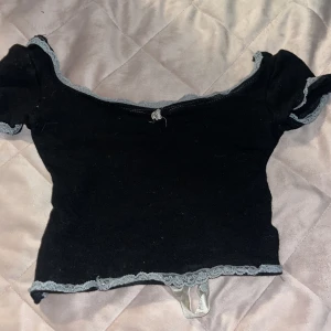 Svart croppad topp med spets och rosett - Supersöt svart croppad topp med ljusgrå spetsdetaljer längs kanterna och en bred sammetsrosett att knyta i ryggen. Toppen   har korta ärmar, perfekt för en trendig och romantisk look.