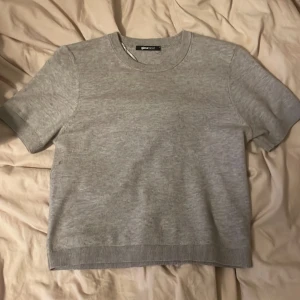 Grå kortärmad tröja Gina Tricot - En basic ljusgrå tröja från Gina Tricot med korta ärmar och rund hals. Tröjan har ribbade muddar vid ärmslut och nederkant, och är gjord i mjuk bomullsmix. Perfekt för en clean och enkel stil.